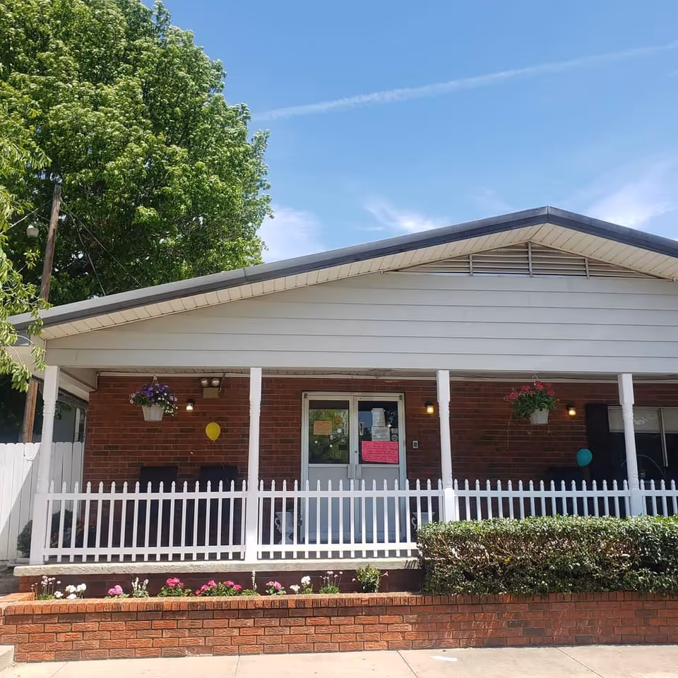 Front exterior view of a single-story brick building with a white picket fence and a covered porch. There are hanging flower pots, a small garden with flowers and bushes, and a clear blue sky above.