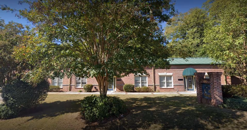 Exterior view of a single-story brick building with multiple windows and two entrances, surrounded by trees and bushes on a sunny day.