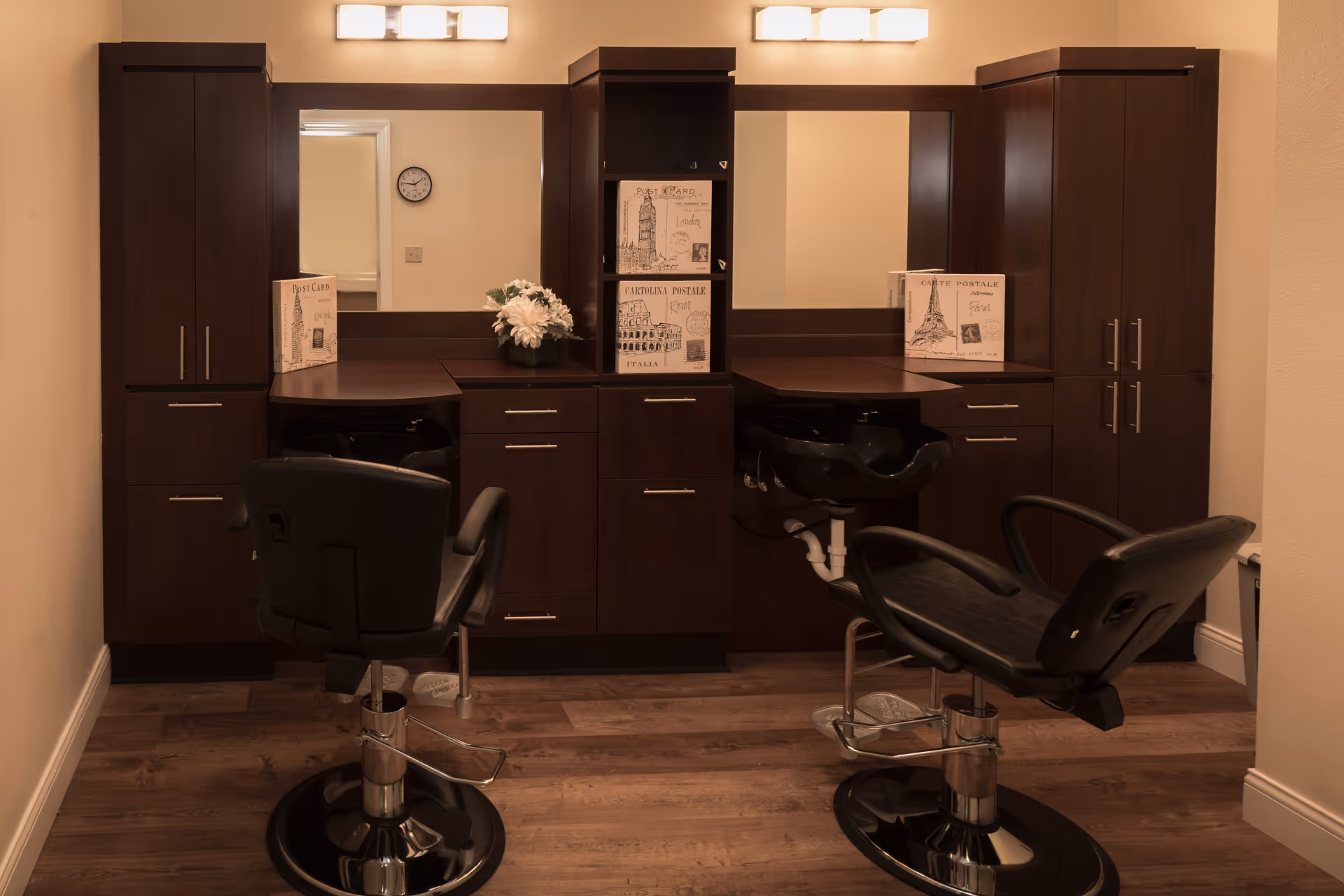 Interior view of a salon area in a senior living facility with two black salon chairs in front of dark wood cabinetry and mirrors. The cabinetry includes shelves with decorative boxes and a small vase with white flowers. The floor is wooden, and there are two light fixtures above the mirrors.