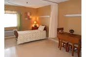 A nursing facility room with a single bed covered with a white patterned blanket, a bedside table with a lamp, a window with green curtains, a small wooden table with two chairs, and a privacy curtain partially drawn.