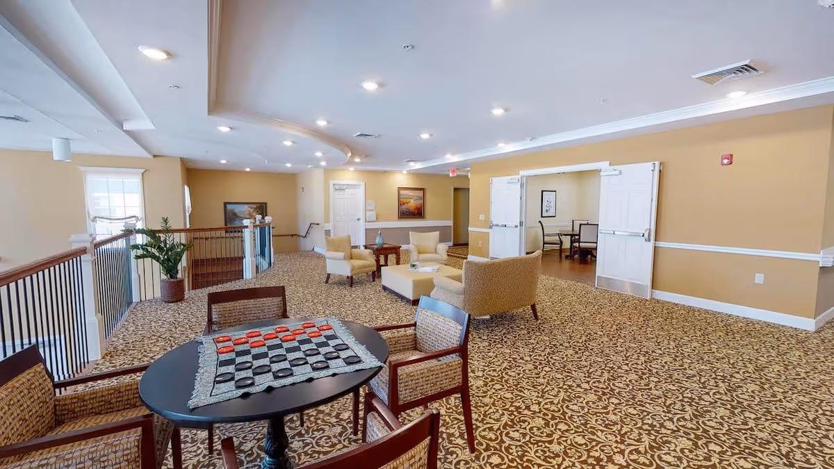A spacious senior living facility common area with patterned carpet, beige walls, and recessed ceiling lights. There is a round table with a checkers game set up and four chairs around it in the foreground. In the background, there are several armchairs and a coffee table arranged for seating. Double doors lead to another room with additional seating and artwork on the walls.