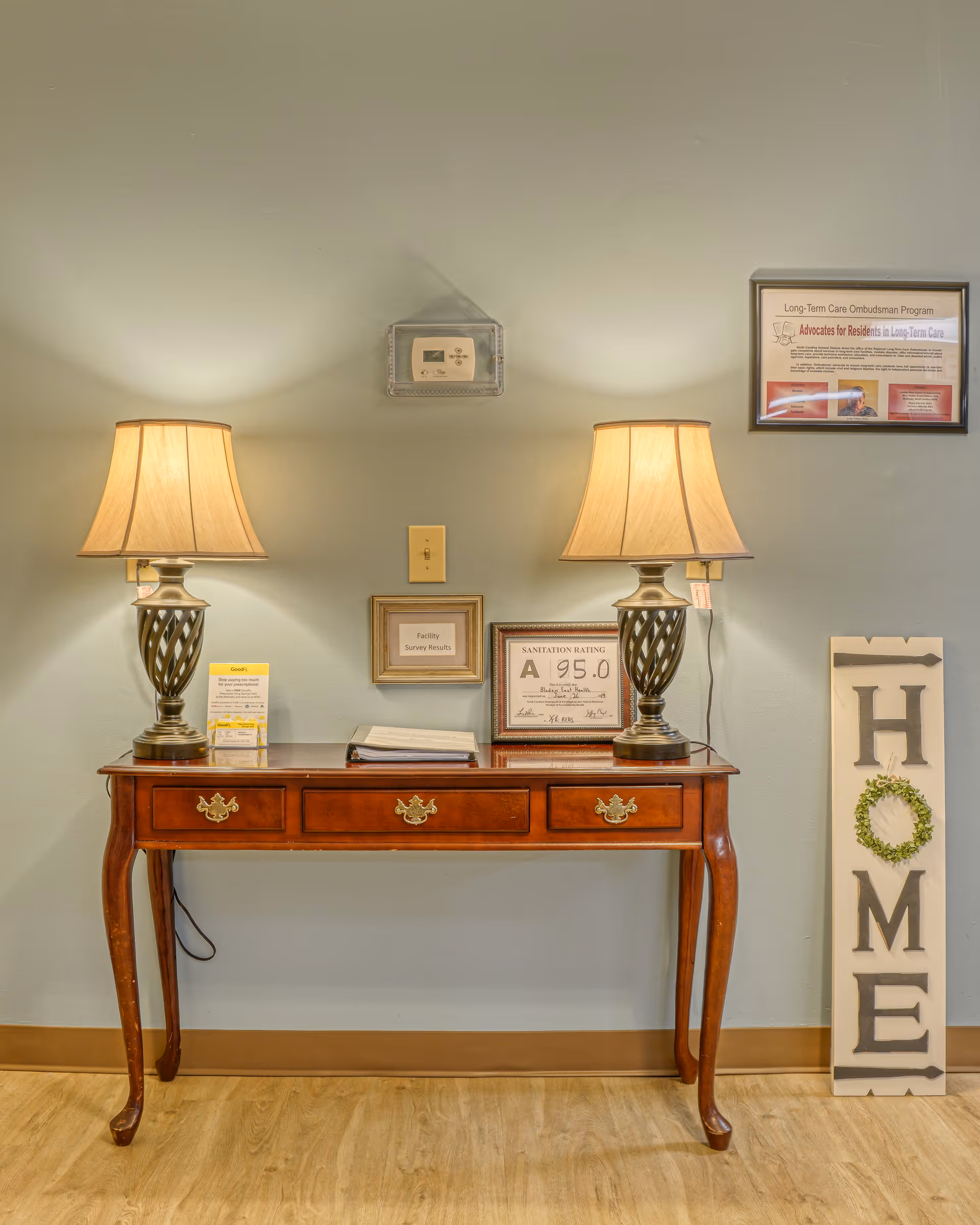 A wooden console table with three drawers is placed against a light green wall. On the table are two decorative lamps with beige shades, a folder, and framed certificates including a sanitation rating of 95.0. Above the table is a thermostat, and to the right on the wall is a framed Long-Term Care Ombudsman Program notice. Leaning against the wall next to the table is a tall decorative sign spelling 'HOME' with a small green wreath replacing the letter O. The floor is light wood with a brown baseboard.