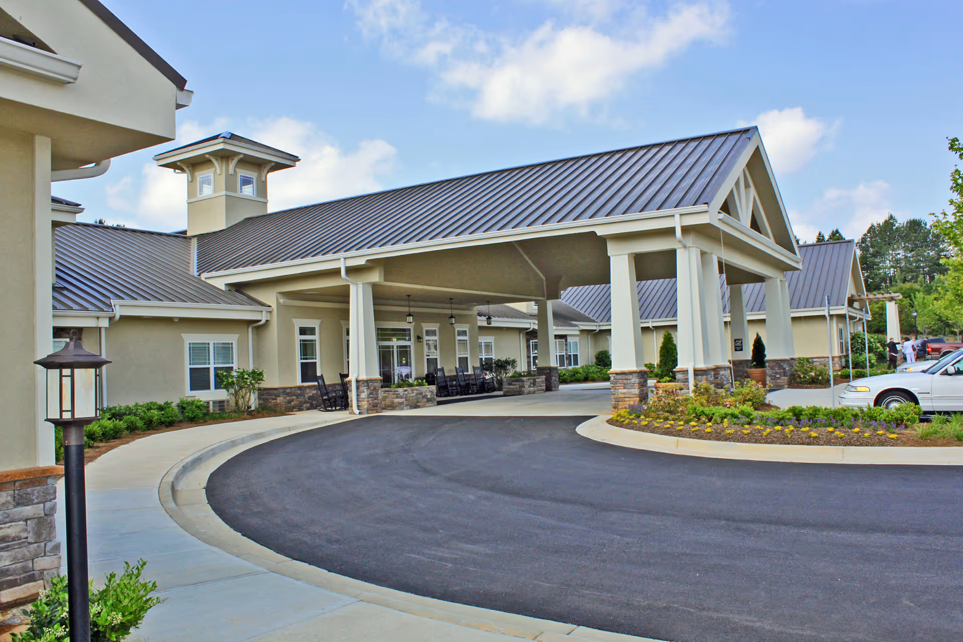 Exterior view of Benton House of Newnan senior living facility showing a covered entrance with stone pillars, a curved driveway, landscaped flower beds, and a few parked cars under a blue sky with some clouds.