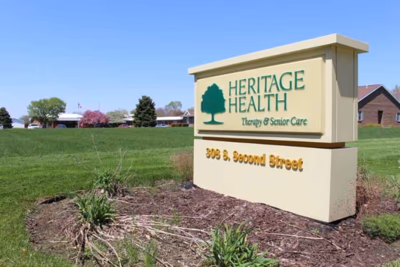 Outdoor view of a beige sign with green and yellow text that reads 'HERITAGE HEALTH Therapy & Senior Care' and '308 S. Second Street' in front of a grassy area with trees and buildings in the background under a clear blue sky.