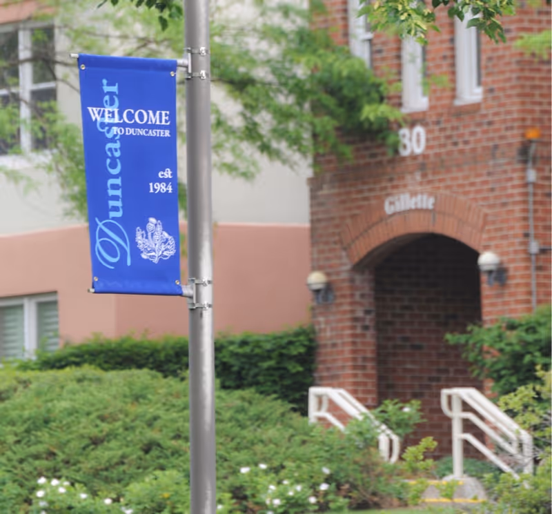 Blue vertical banner on a pole that reads 'Welcome to Duncaster est 1984' with a decorative emblem below the text. In the background, there is a brick building entrance with the number 30 and the name 'Gillette' above the doorway, surrounded by greenery and bushes.