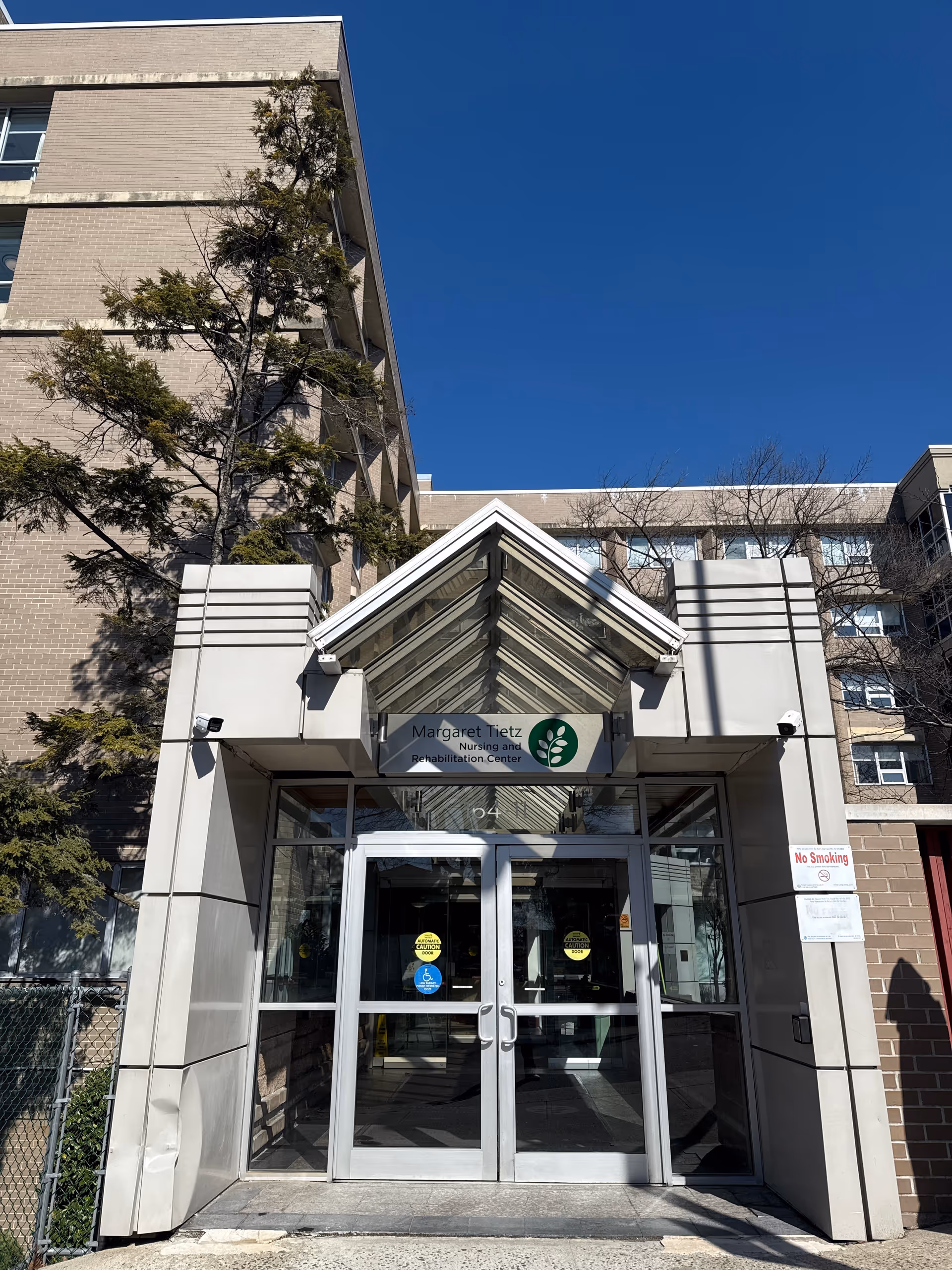 Entrance of Margaret Tietz Nursing and Rehabilitation Center with glass double doors under a glass canopy, beige brick building facade, and a clear blue sky.