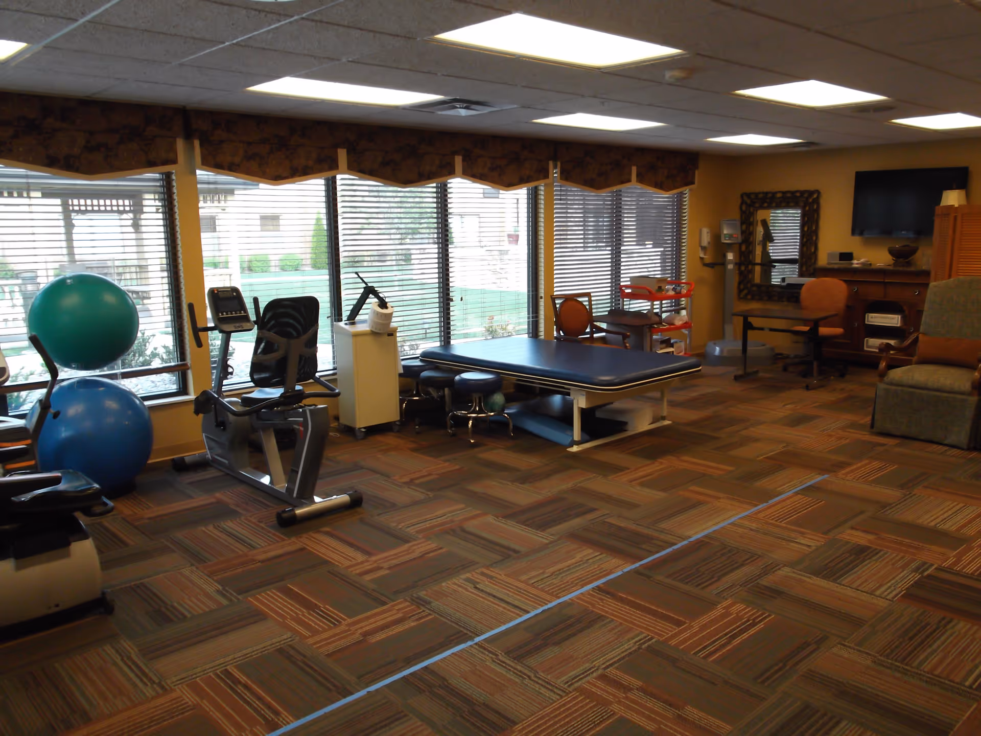 Bright therapy and exercise room with a recumbent bike, treatment table, large exercise balls and seating by large windows.
