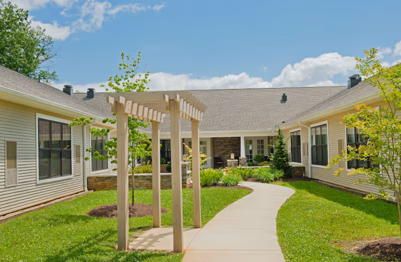A sunny outdoor courtyard area at Asbury Place Maryville featuring a paved walkway, green grass, small trees, and a wooden pergola structure. The courtyard is surrounded by single-story beige buildings with multiple windows under a blue sky with some clouds.