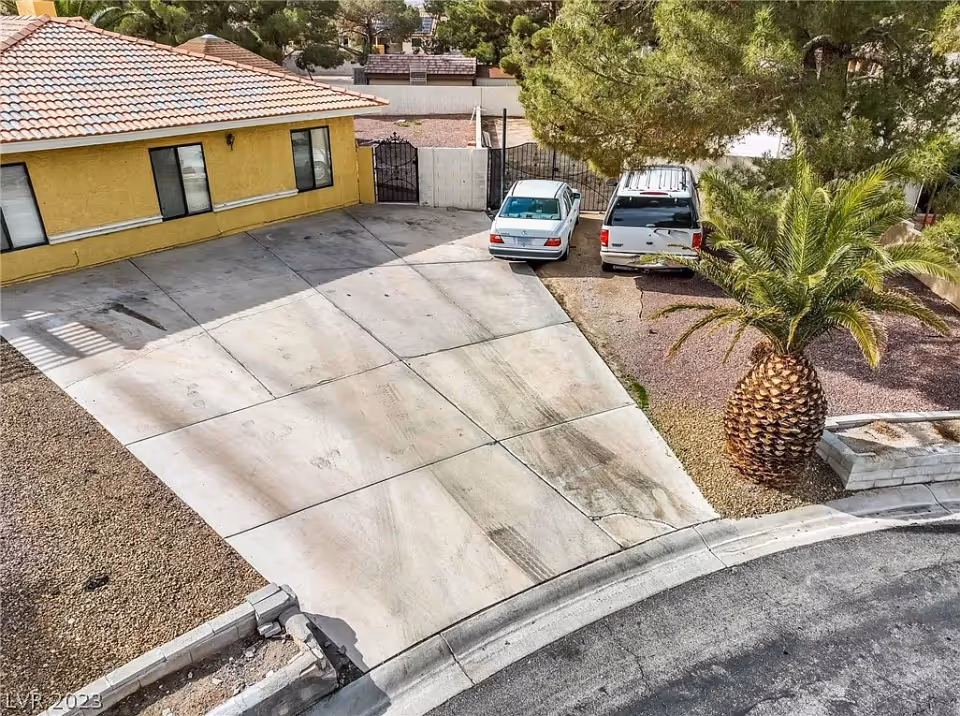 A driveway with two parked cars next to a yellow building with a tiled roof. There is a palm tree and some other trees on the right side, and a gated fence in the background.