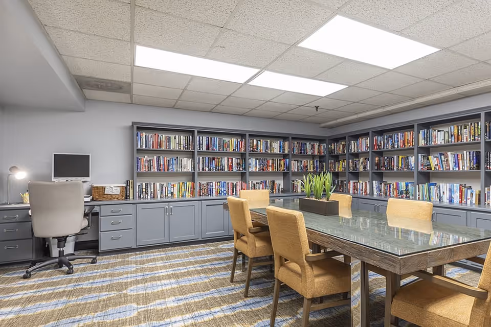 A well-lit reading/library room with wall-to-wall bookshelves, a glass-top table surrounded by upholstered chairs, and a small desk with a computer.