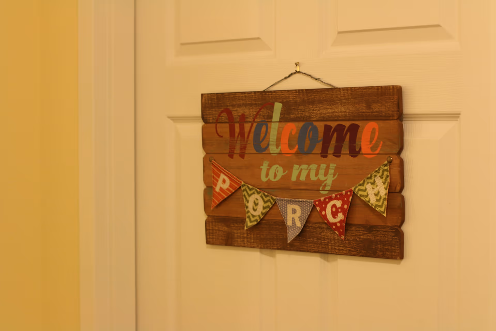 A wooden sign hanging on a white door with the colorful text 'Welcome to my porch' and small decorative flags spelling 'PORCH'.