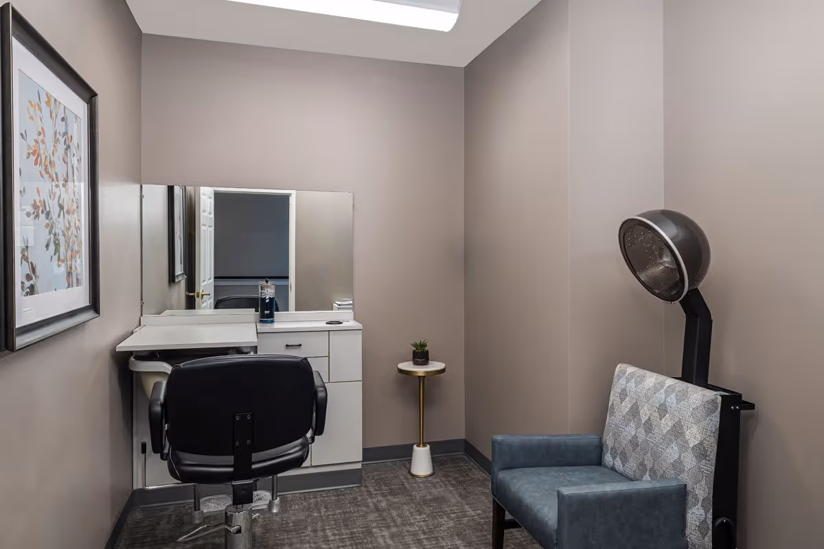 A small salon room with a black salon chair in front of a white vanity with a mirror. There is a framed artwork on the left wall, a small round side table with a small plant, and a blue patterned chair with a hair dryer hood attached behind it. The walls are painted beige and the floor is carpeted.