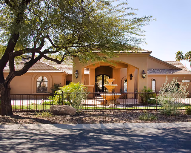 Front exterior of a single-story stucco building with an arched entrance, tiered fountain, tree and low metal fence.