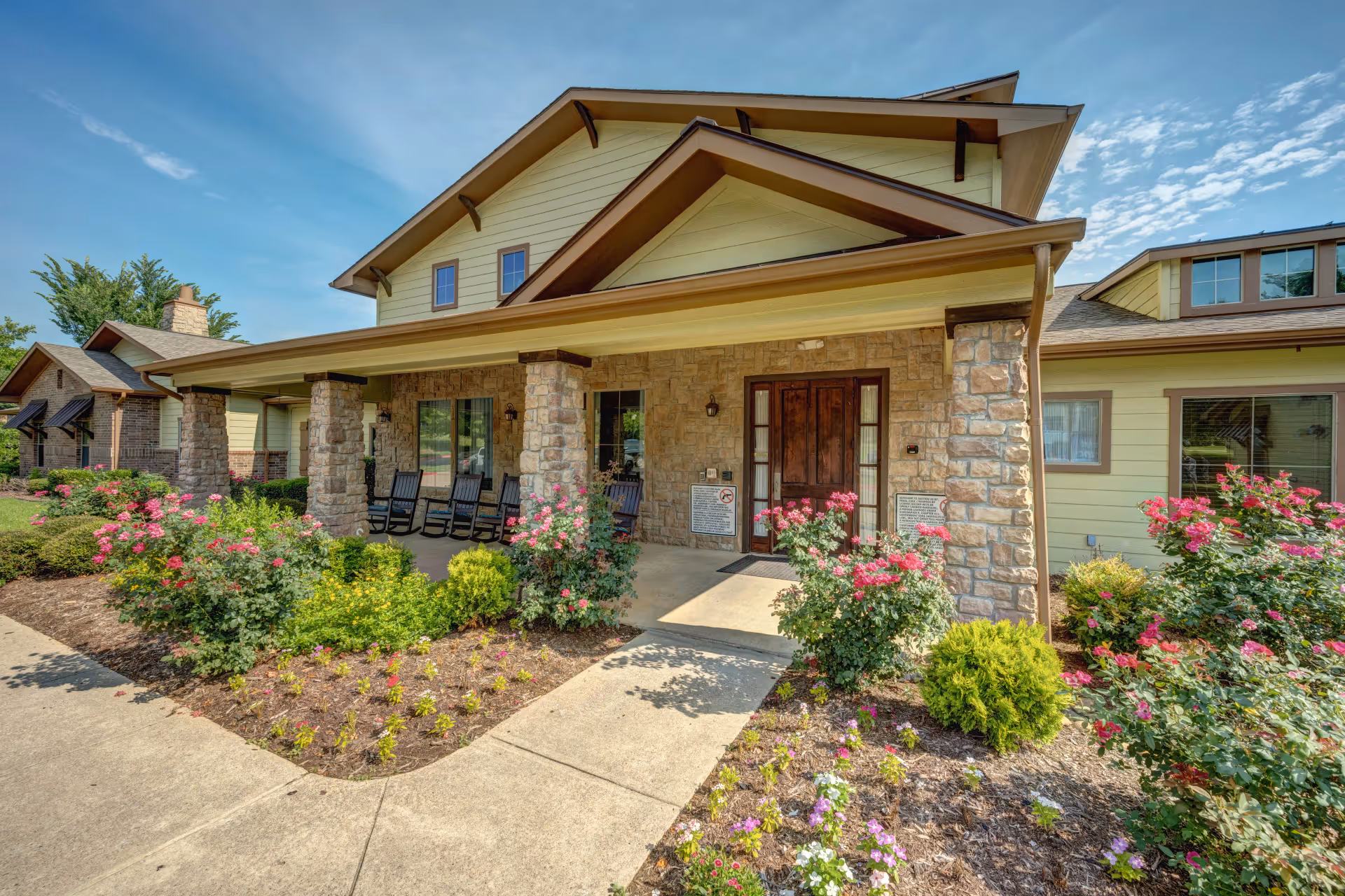 Front exterior view of Hawkins Creek Assisted Living and Memory Care facility featuring a covered porch with stone pillars, wooden rocking chairs, a wooden front door, and landscaped flower beds with pink and purple flowers under a clear blue sky.