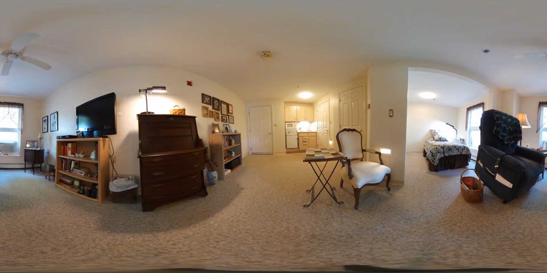 A panoramic view of a senior living community apartment showing a living area with a TV on a wooden shelf, a dark wooden dresser, framed pictures on the wall, a small kitchen with cabinets and appliances, a white upholstered chair with a small table, a bedroom area with a bed and windows, and a recliner chair with a basket beside it. The room is carpeted and well-lit with natural light from the windows.