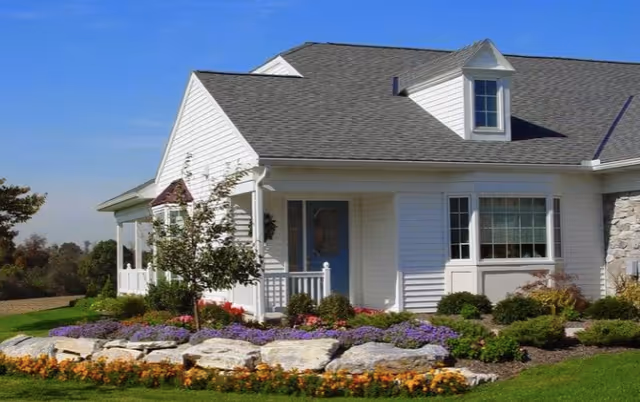 A white single-story house with a gray shingled roof, a small front porch with a blue door, and a dormer window. The house is surrounded by a well-maintained garden with colorful flowers, shrubs, and rocks under a clear blue sky.