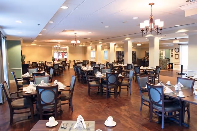 A spacious dining room with multiple wooden tables and chairs arranged neatly. Each table is set with white cups, saucers, napkins, and cutlery. The room has warm lighting with chandeliers hanging from the ceiling and a wooden floor. The walls are painted in soft yellow and beige tones, and there are columns and decorative elements throughout the space.