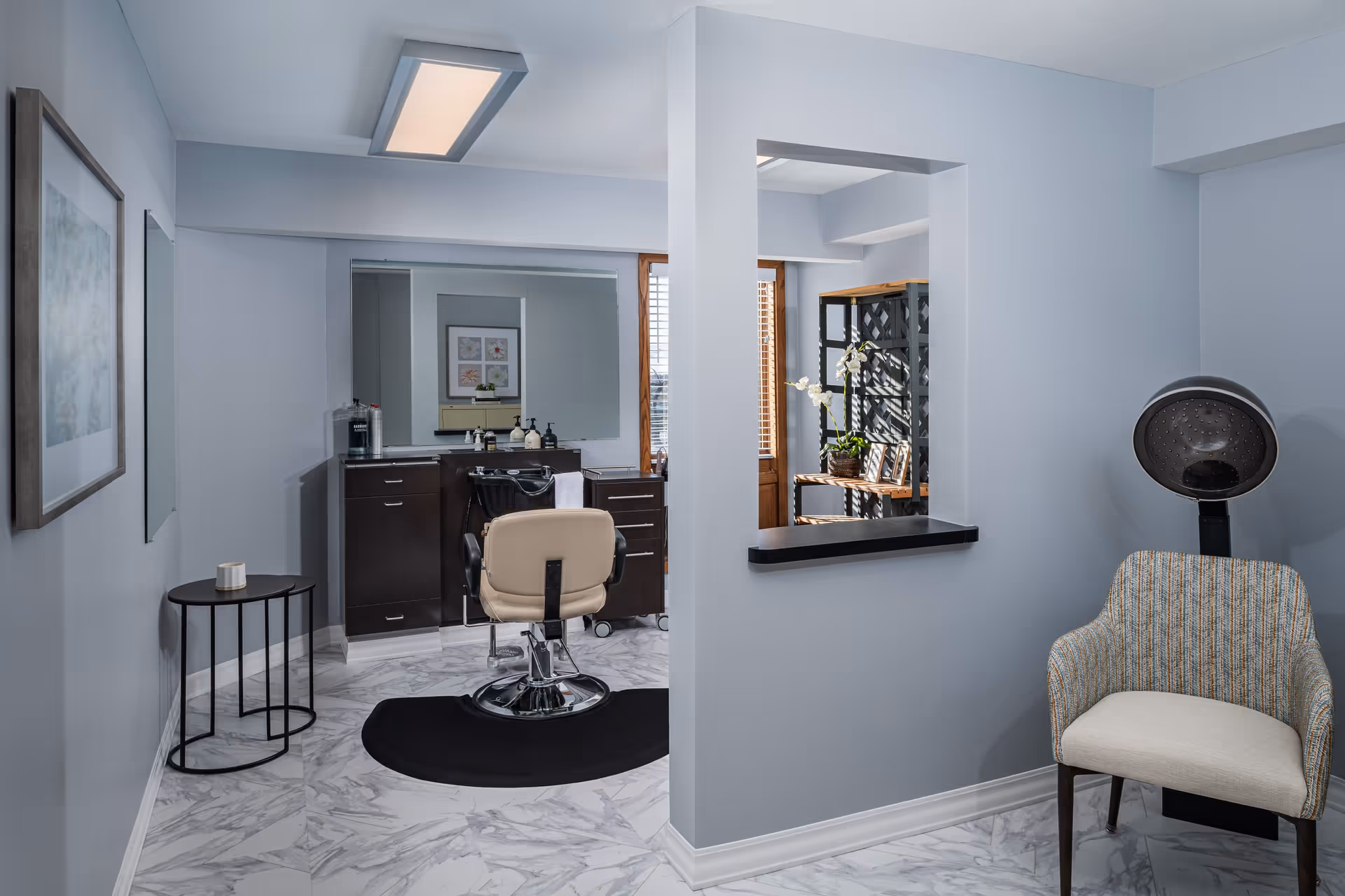 Small salon-style interior with a barber chair, styling station, hooded hair dryer and seating in a light-blue room.