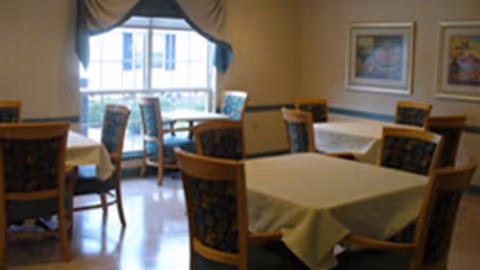 A dining room with several square tables covered with beige tablecloths and surrounded by wooden chairs with patterned cushions. There is a large window with draped curtains allowing natural light to enter the room. Two framed pictures hang on the wall.