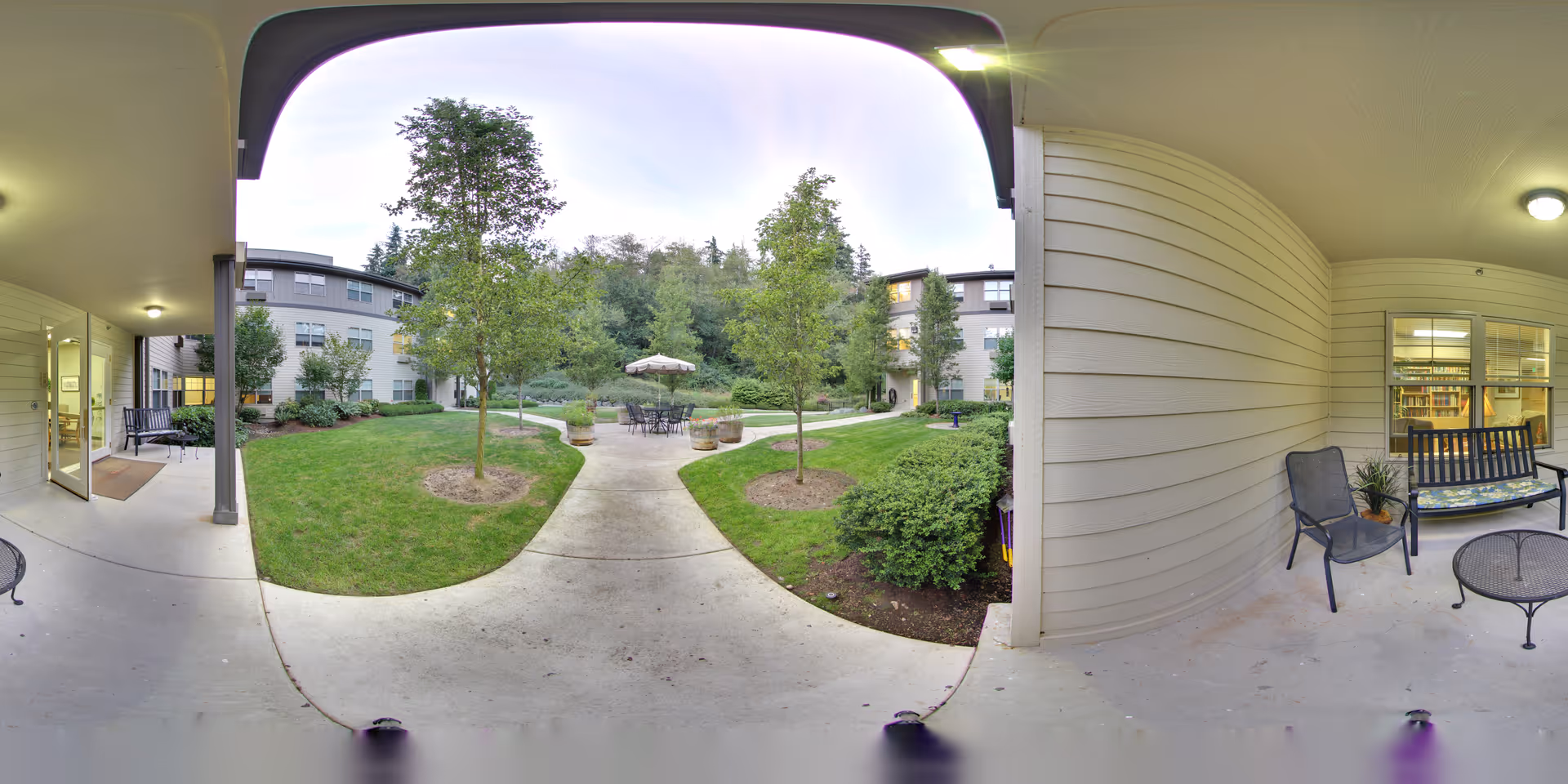 Covered patio and walkway opening onto a landscaped courtyard with trees, outdoor seating and surrounding apartment building.