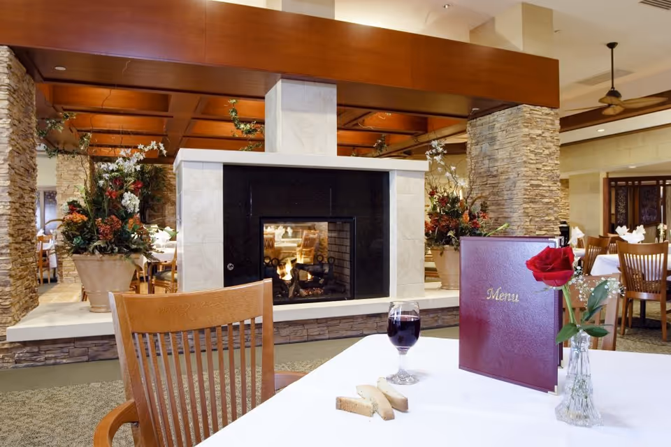 Dining room with a central double-sided fireplace, wooden chairs and tables, and a foreground table holding a menu, glass of red wine, a rose, and bread.