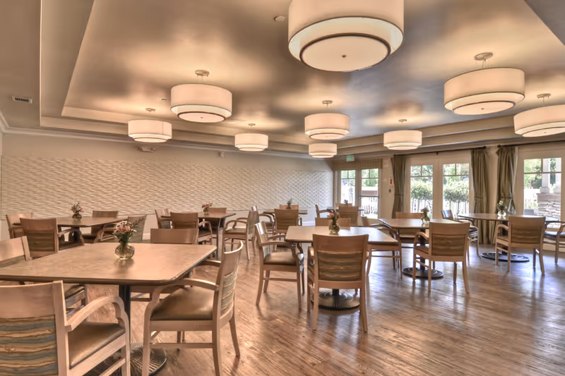 A spacious dining room with multiple square and round tables surrounded by wooden chairs. Each table has a small vase with flowers. The room features large windows with green curtains allowing natural light to enter. The ceiling has several modern circular light fixtures, and the floor is made of wood.