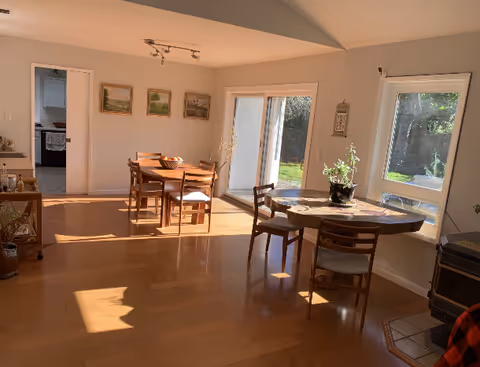 Sunlit dining area with two wooden tables and chairs on hardwood floors, sliding glass door and windows overlooking a backyard.