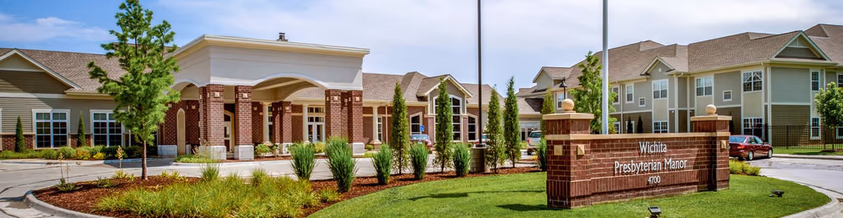 Front exterior view of Wichita Presbyterian Manor, showing a large brick and beige building with a covered entrance, landscaped greenery, and a sign with the facility name and address in front.