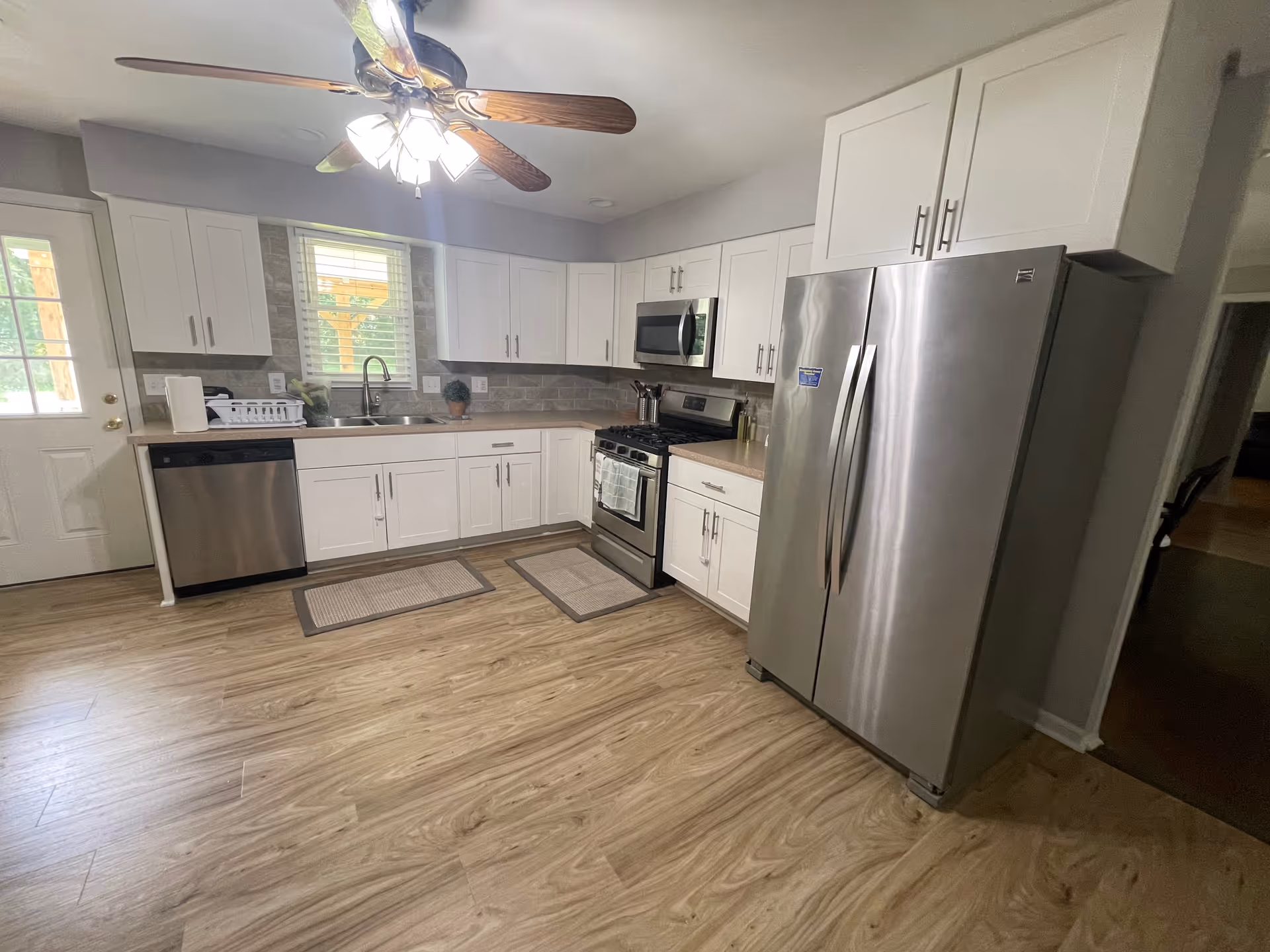 Bright modern kitchen with white cabinets, stainless steel refrigerator and stove, a sink under a window, and wood-look flooring.