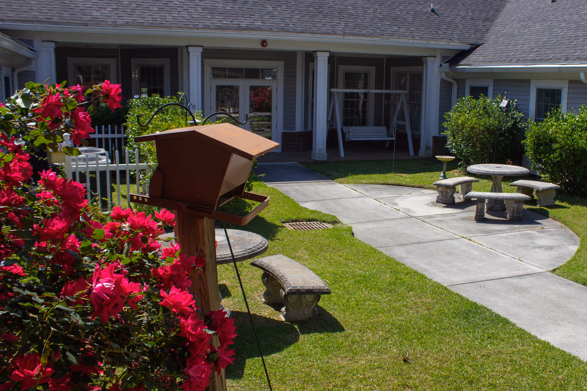 Outdoor courtyard area with a concrete pathway, green grass, and bright pink flowers in the foreground. There is a bird feeder mounted on a wooden post, stone benches and a round stone table. In the background, there is a building with a covered porch and a white swing.