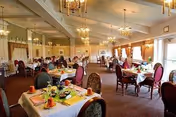 Spacious dining room with multiple set tables, upholstered chairs, chandeliers, and people seated at some tables.