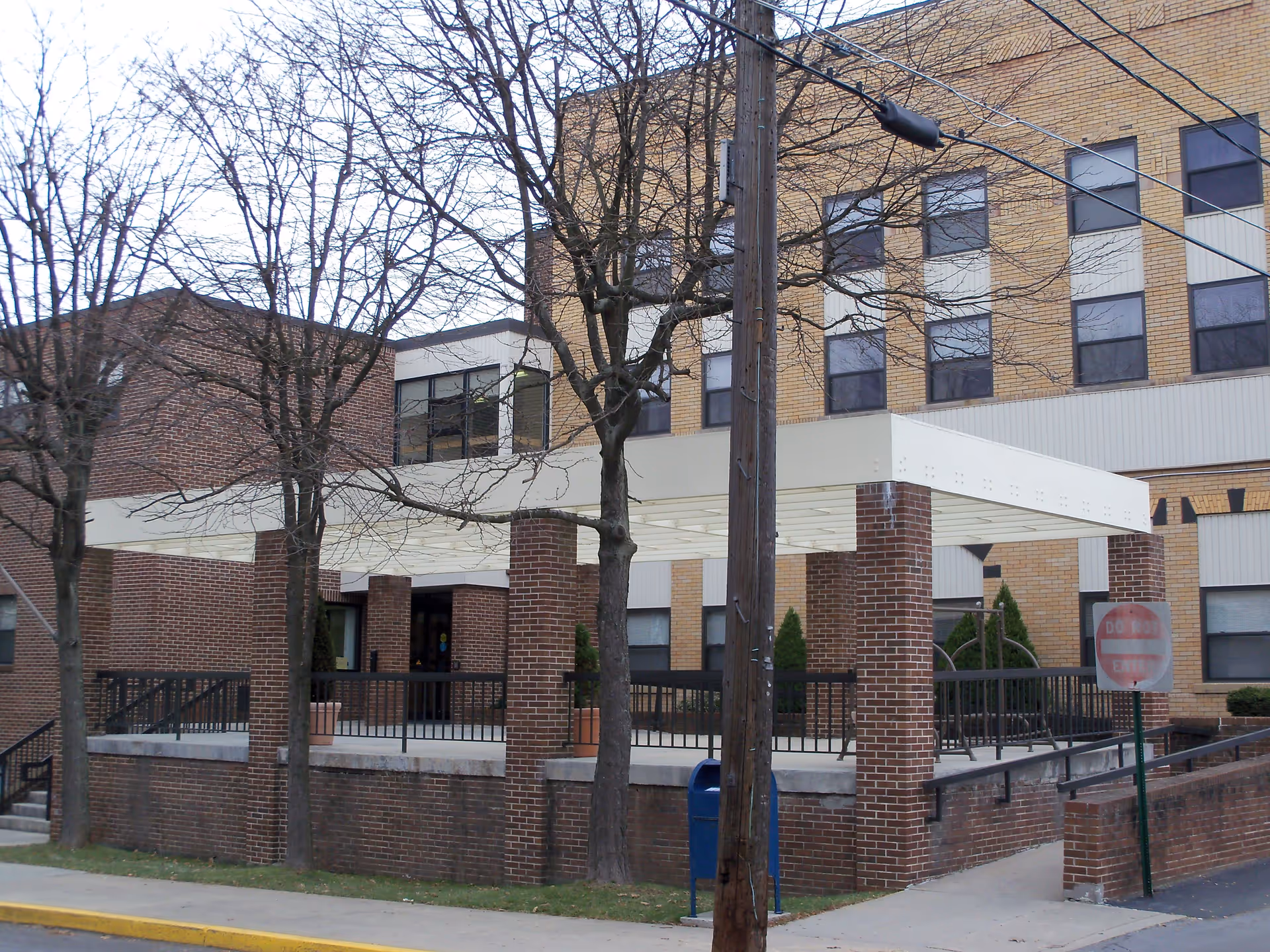 Exterior view of a multi-story brick building with a covered entrance supported by brick pillars. Leafless trees and a utility pole are visible in front of the building, along with a blue mailbox and a 'Do Not Enter' sign near the sidewalk.