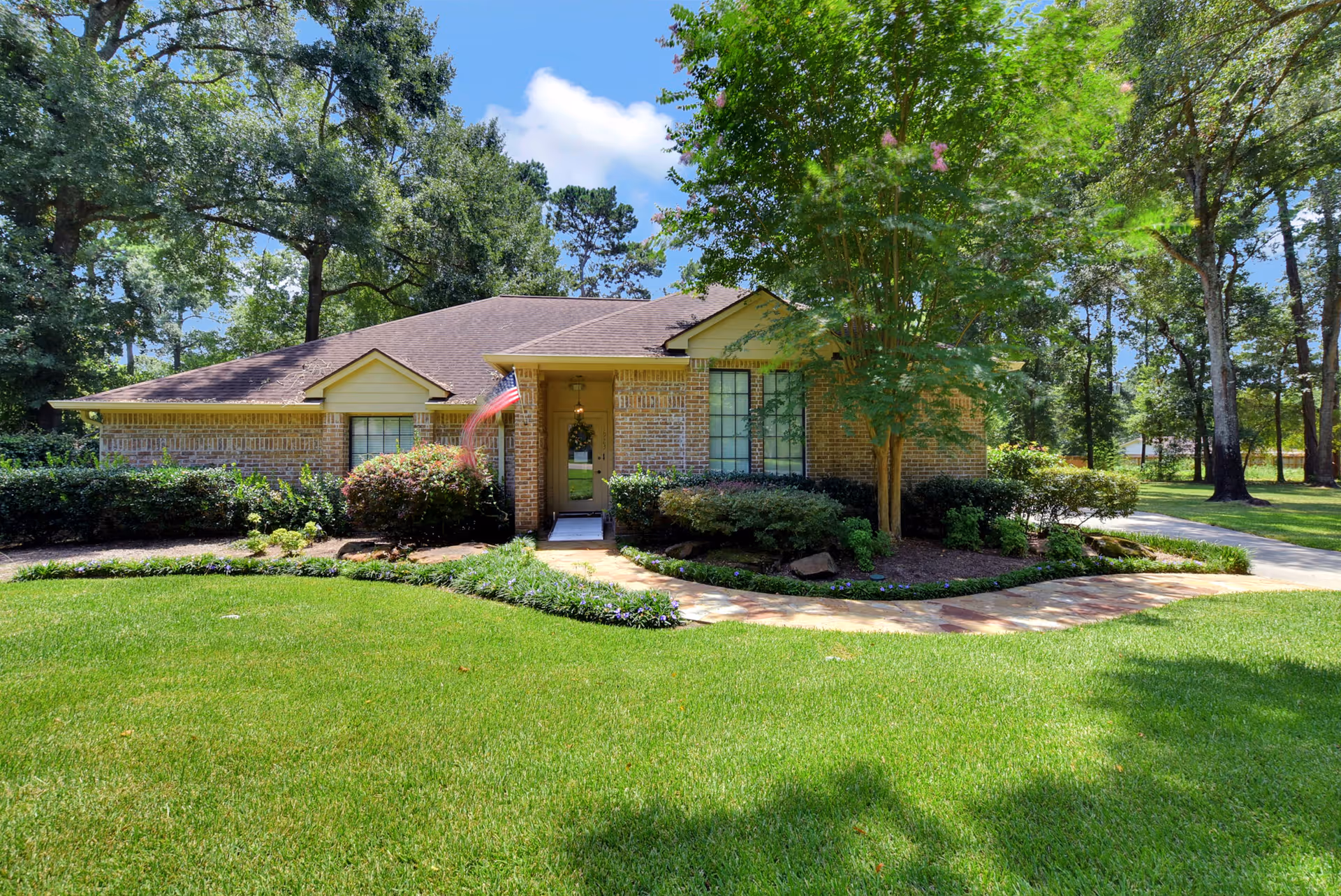Single-story brick house with a brown roof surrounded by green grass, shrubs, and trees under a blue sky with some clouds. A stone pathway leads to the front door, which has an American flag hanging beside it.
