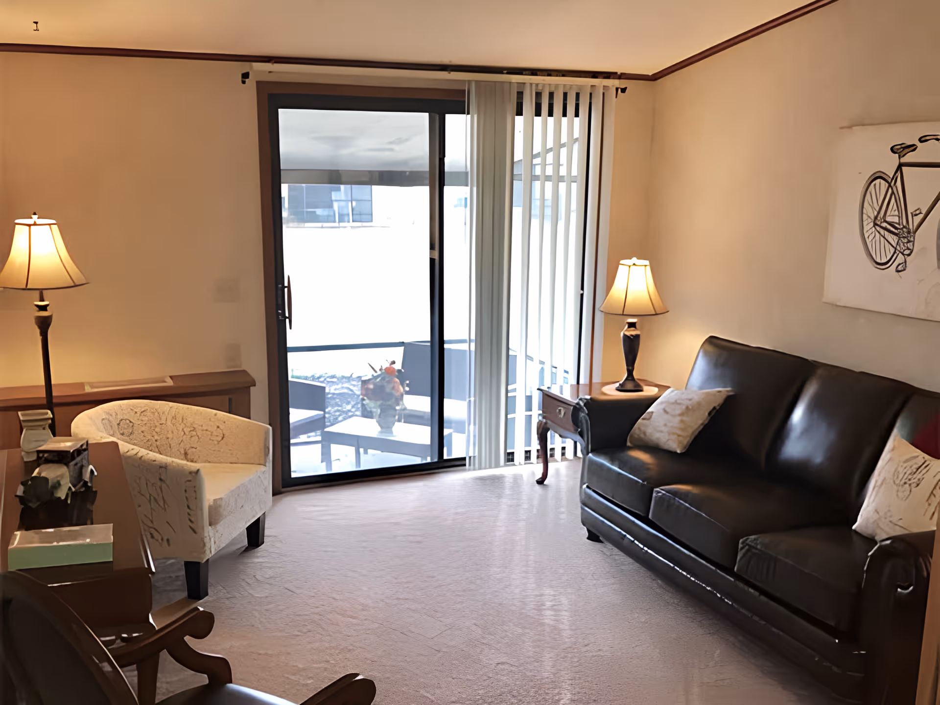 Bright living room with a leather sofa, armchair, side tables and lamps facing a sliding glass door to a small patio.