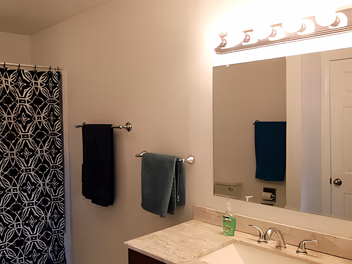 A bathroom with a marble countertop sink, a large mirror above it, two towel racks with dark-colored towels, and a shower with a black and white patterned curtain.