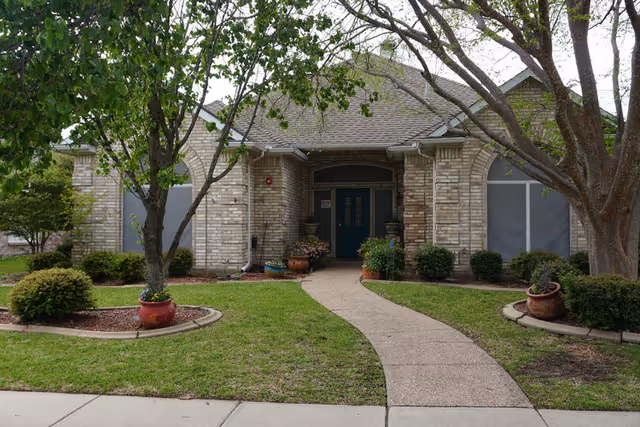 Front exterior of a single-story brick house with a curved walkway, trees, and potted plants leading to a blue double front door.