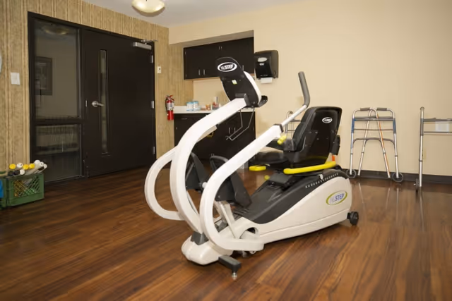 A rehabilitation center room with a recumbent exercise bike in the center, a walker and a rollator against the wall, a fire extinguisher mounted near a black door, and a small counter with a paper towel dispenser and some supplies.