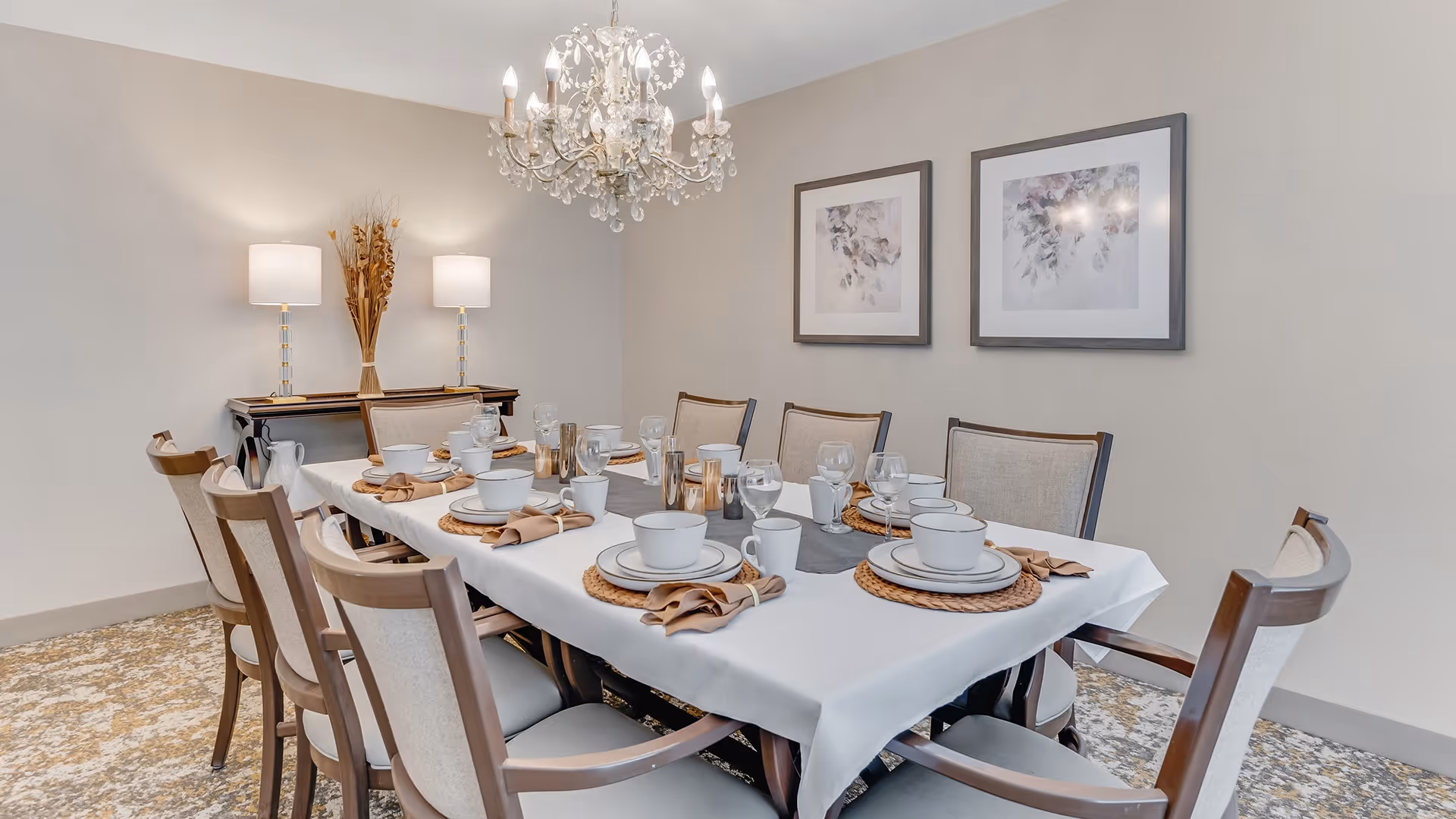 A formal dining room with a long rectangular table set for eight people. The table is covered with a white tablecloth and has place settings including plates, bowls, cups, glasses, and napkins on woven placemats. The room features a chandelier hanging above the table, two table lamps on a sideboard against the wall, and two framed floral artworks on the wall.