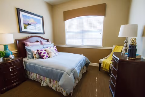 A neatly made bed with light blue bedding and multiple decorative pillows in a bedroom. The room has beige walls, a window with blinds and a brown valance, a wooden nightstand with a lamp and a clock, a wooden dresser with a lamp and decorative items, and a chair with a yellow throw blanket.