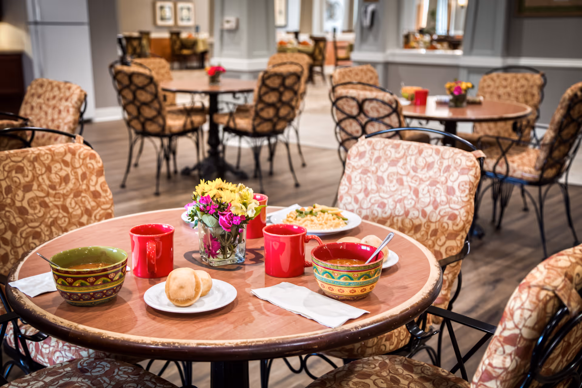 A dining room table in an assisted living facility set with soup bowls, red mugs, a bread roll, and a small vase of flowers with other tables and chairs in the background.