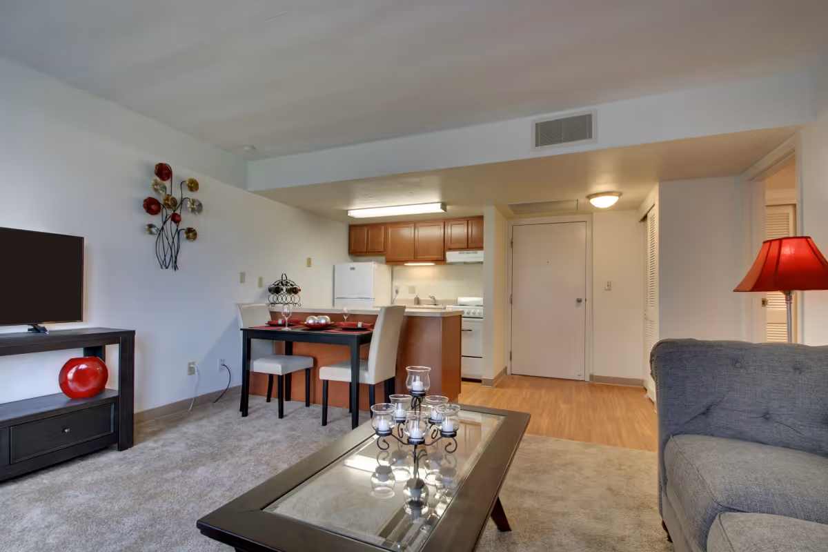 Interior view of a senior living apartment featuring a living room with a gray couch, glass coffee table with candle holder, and a TV on a stand. Adjacent to the living room is a dining area with a small table set for two and a kitchen with wooden cabinets, a refrigerator, and a stove. The space has carpeted and wood flooring with neutral-colored walls and ceiling lights.
