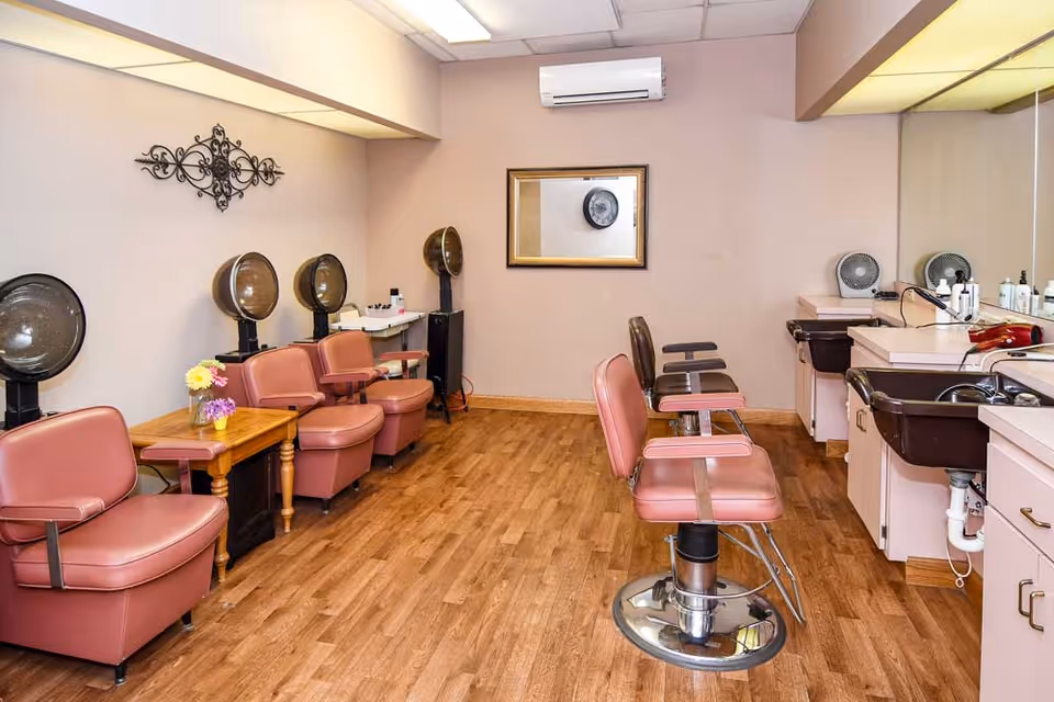 Interior view of a salon area with pink salon chairs, hair dryers, a wooden side table with flowers, mirrors, and sinks for washing hair. The room has wooden flooring and beige walls with a decorative wall piece and a framed mirror.
