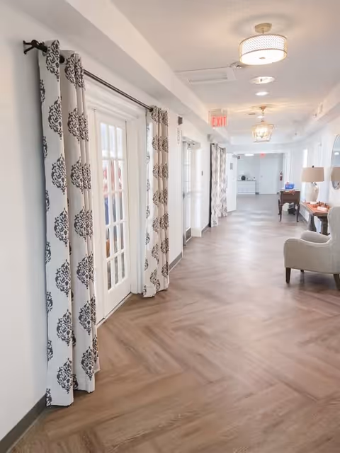 A bright hallway with wood-patterned flooring and white walls. The hallway features multiple glass-paneled doors with patterned curtains, ceiling light fixtures, an exit sign, and some furniture including a chair and a table with a lamp along the right side.