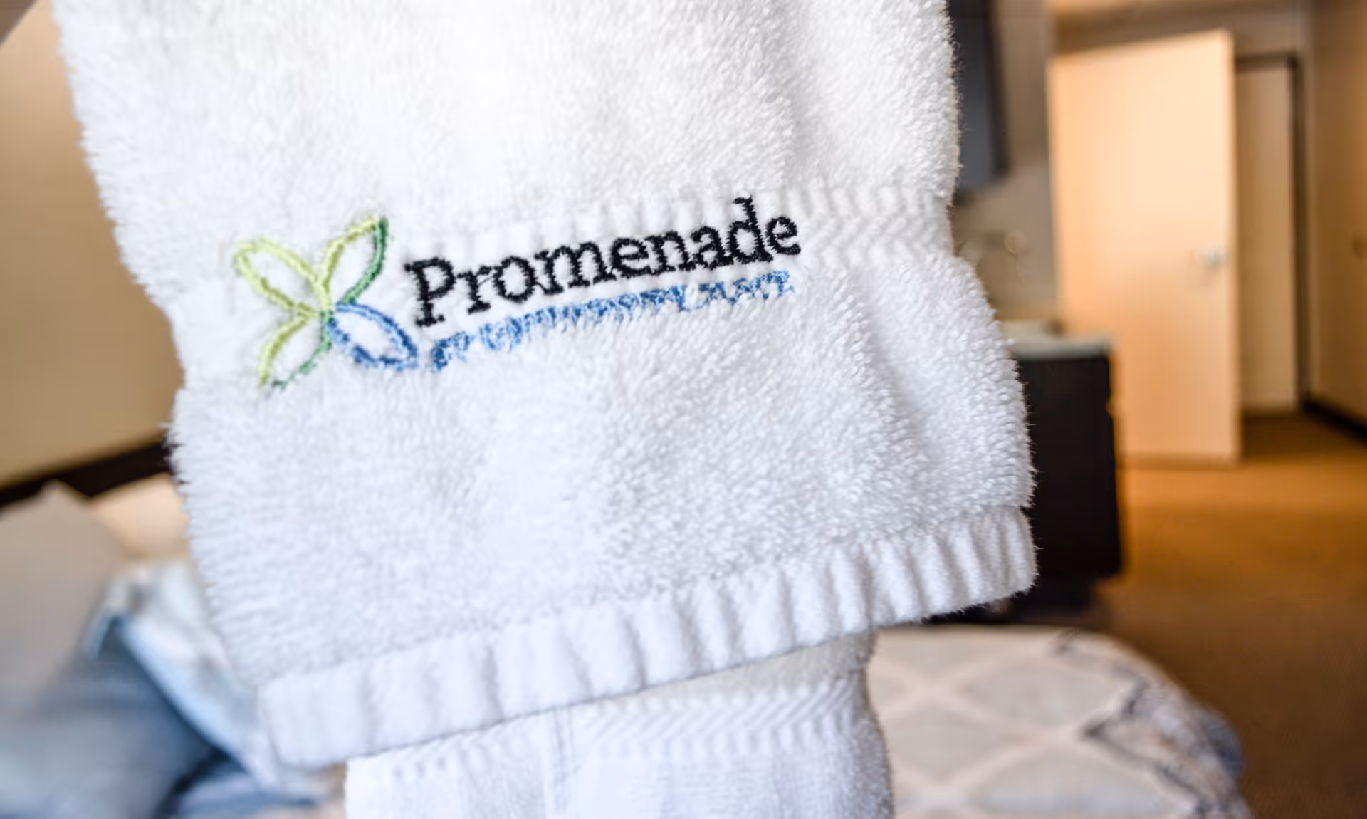 Close-up of a white towel embroidered with the logo and name 'Promenade at University Place' in a softly lit room with a bed and an open door in the background.