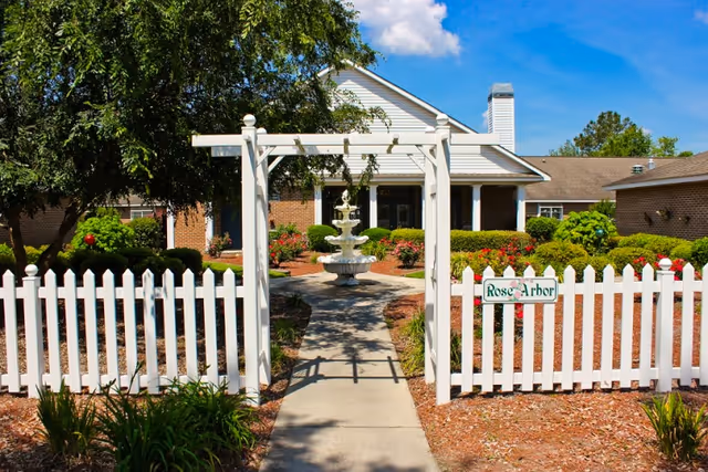 A white picket fence with an arbor entrance leading to a concrete pathway. The pathway goes past a tiered water fountain and is surrounded by well-maintained bushes and flowers. In the background, there is a brick building with a white porch and chimney under a clear blue sky.