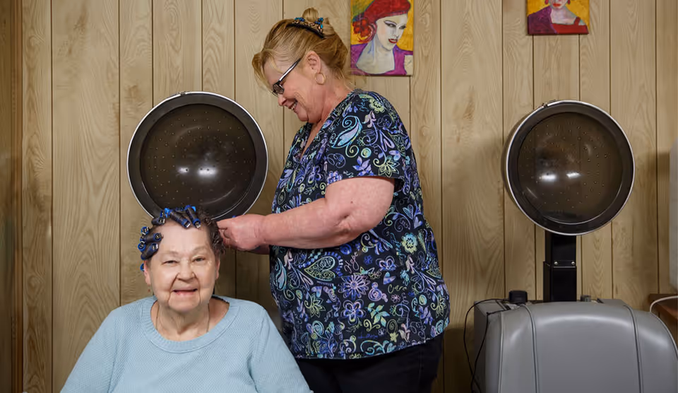 An elderly woman with hair rollers sits smiling while a hairdresser stands behind her, styling her hair under a hair dryer in a room with wood-paneled walls and colorful portraits on the wall.