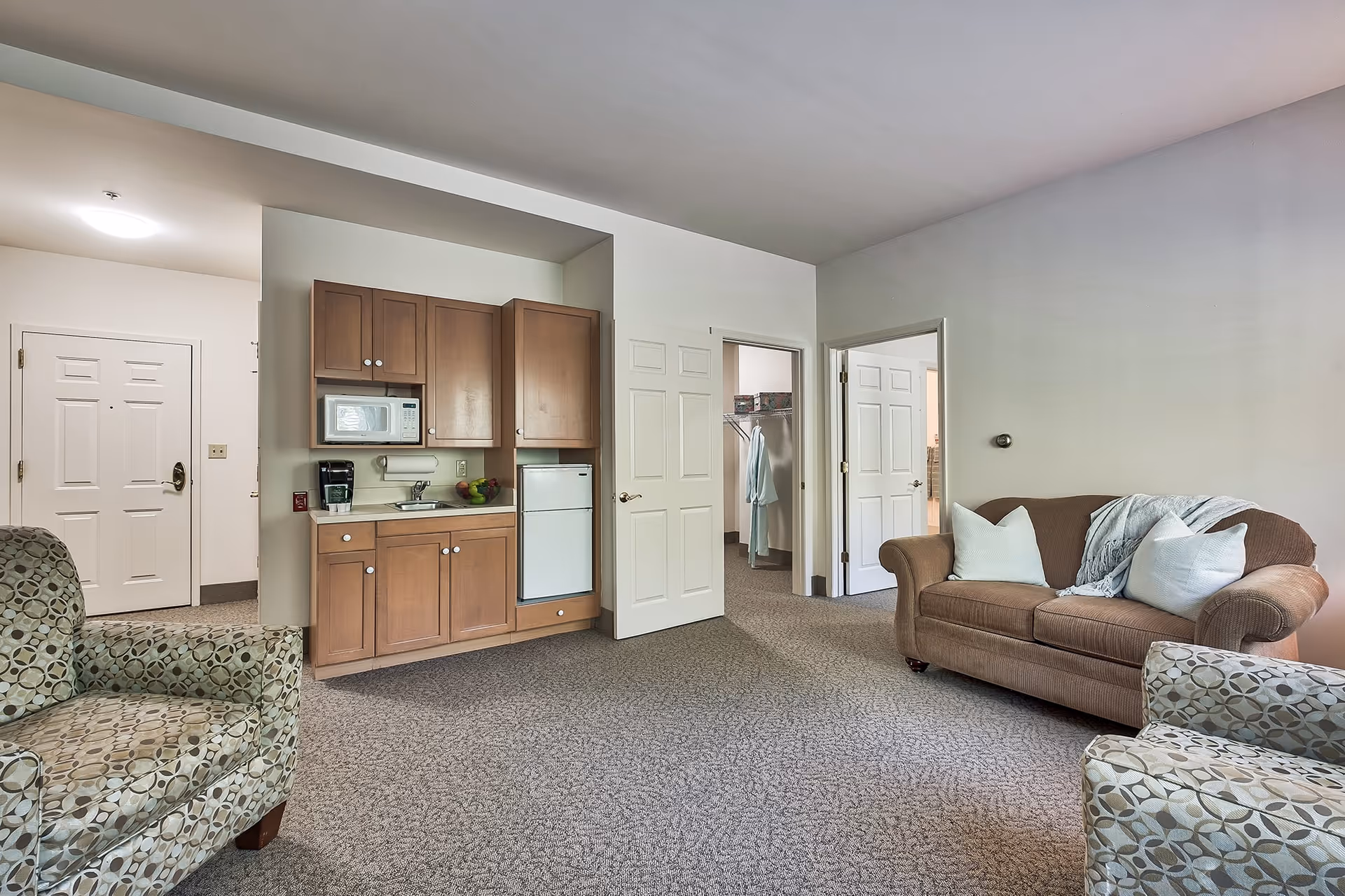 A cozy living room area in a senior living facility with a brown couch adorned with two white pillows and a gray throw blanket, two patterned armchairs, a small kitchenette with wooden cabinets, a microwave, a coffee maker, and a mini fridge. There are two open doors leading to other rooms, one showing a closet with hanging robes. The walls are light-colored and the floor is carpeted.