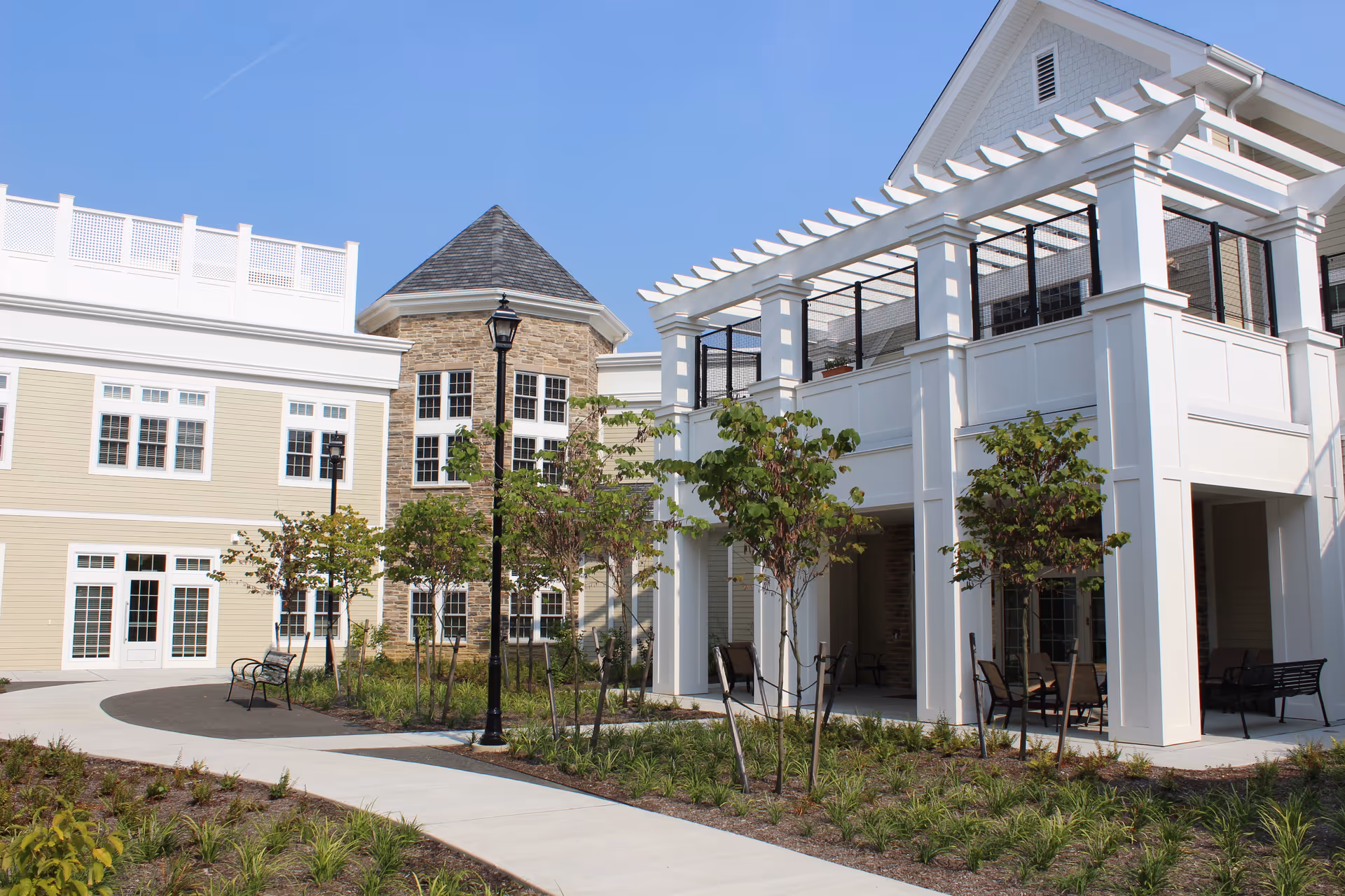 Outdoor view of a senior living facility named Parker at Monroe featuring a landscaped garden with young trees, benches, a paved walkway, and a building with a stone tower and white pergola structure under a clear blue sky.