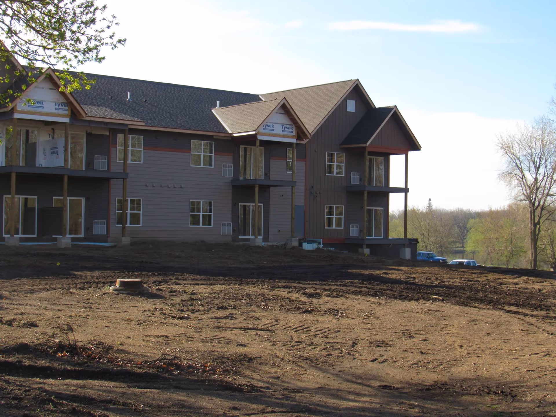 Exterior view of a two-story building under construction with brown siding and multiple windows, surrounded by bare soil and some trees in the background under a clear sky.