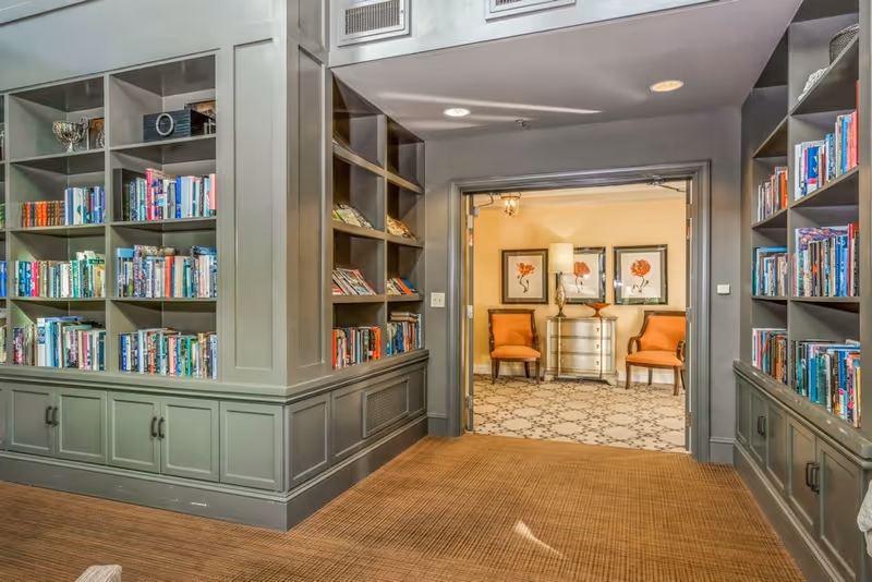 Interior view of a library or reading area with built-in bookshelves filled with books on both sides. The shelves are painted gray. Through an open doorway, a sitting area is visible with two orange upholstered chairs, a silver chest of drawers, a table lamp, and three framed floral artworks on the wall.